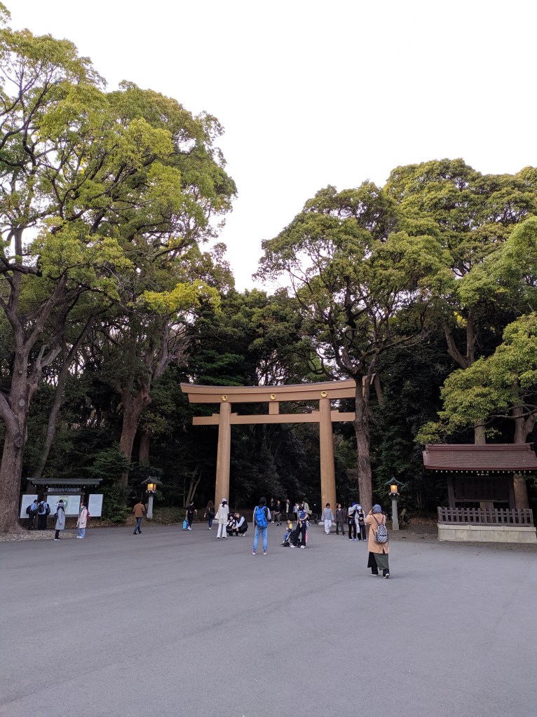 Pathway that leads to a big fair wooden torii from the Meiji Jingu Sanctuary. Large trees with green leaves are on the back of the torii hiding the pathway to the inside of the sanctuary park. People can be seen walking in the direction of the torii and passing through it.