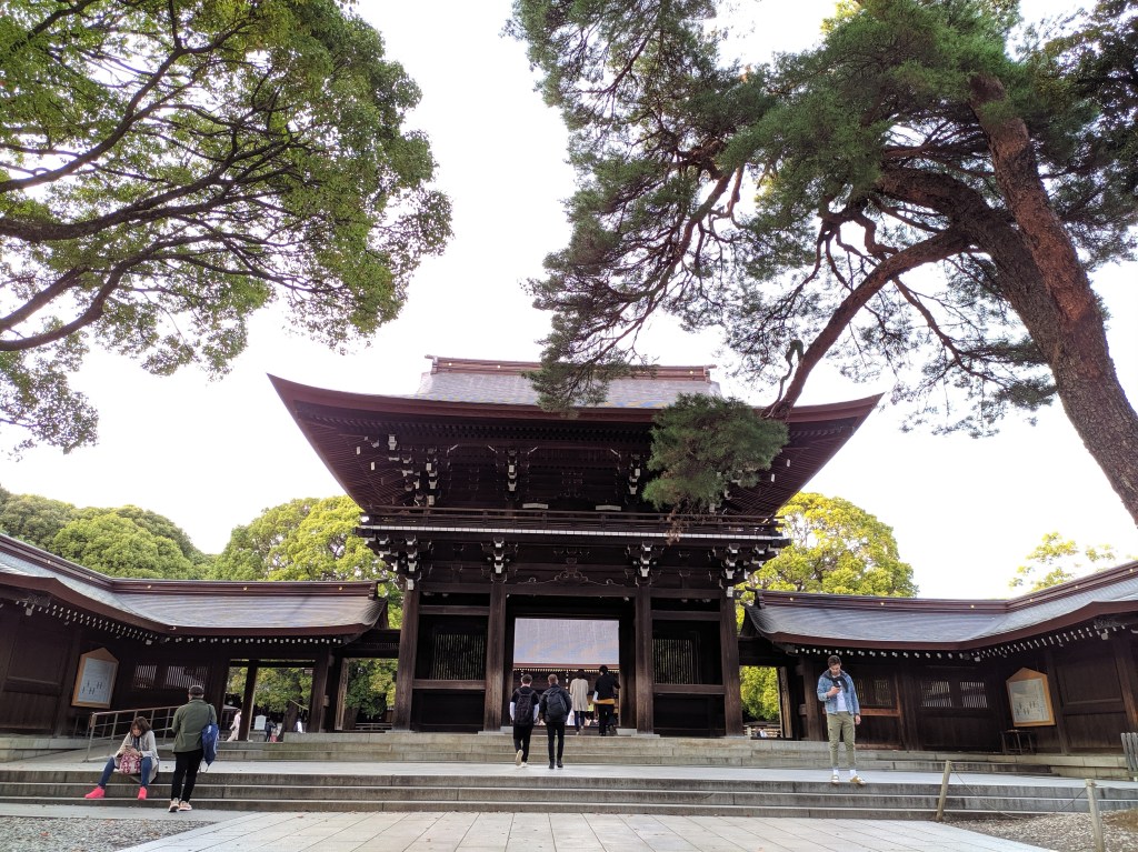 Vista do santuário Meiji Jingu em Tóquio, com estruturas de madeira e visitantes ao redor, cercado por árvores verdes.