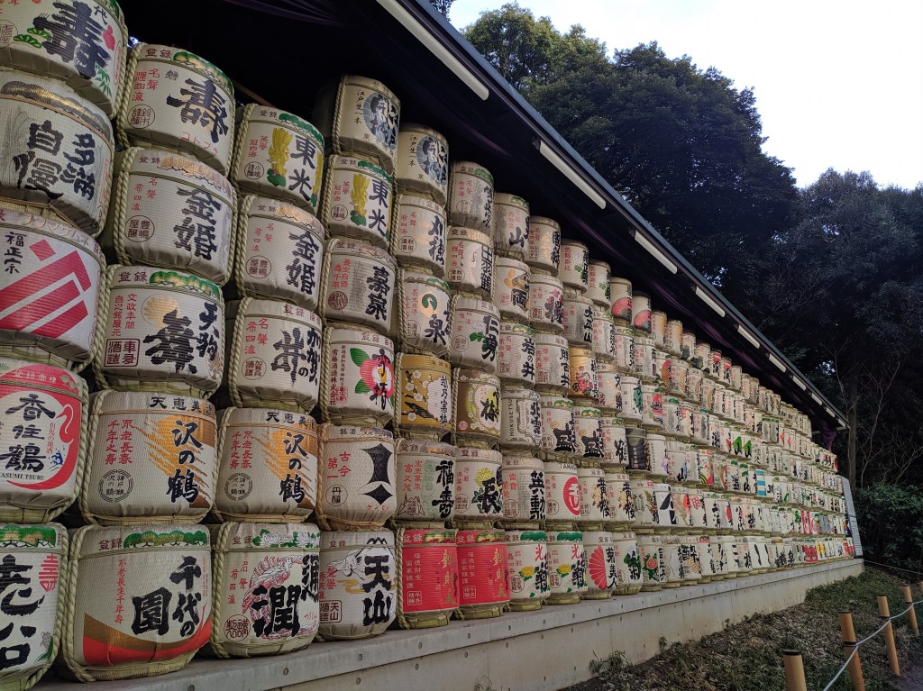Vários barris de saquê decorando uma parede, com rótulos coloridos e desenhos variados, cercados por árvores ao fundo.